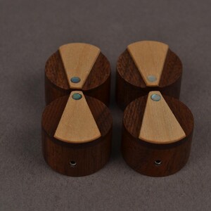 Puede incluir: Cuatro pomos cil&iacute;ndricos de madera marr&oacute;n oscuro, cada uno con una incrustaci&oacute;n triangular de madera beige claro en la superficie superior. Un peque&ntilde;o acento de piedra azul verdosa iridiscente est&aacute; incrustado en la punta de cada tri&aacute;ngulo.