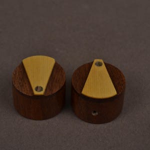 Pu&ograve; includere: Due manopole cilindriche realizzate in legno marrone scuro, ciascuna caratterizzata da un inserto triangolare in legno beige chiaro sulla superficie superiore. Ogni manopola presenta un piccolo foro circolare all'estremit&agrave; stretta dell'inserto.