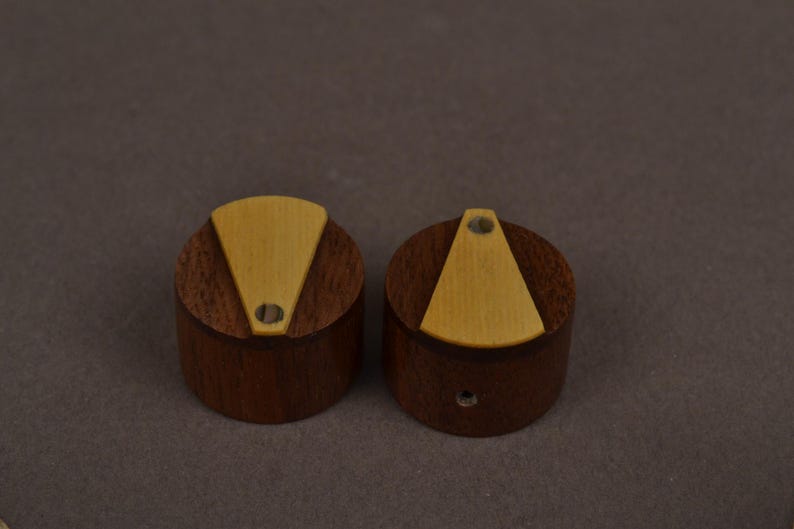 Pu&ograve; includere: Due manopole cilindriche realizzate in legno marrone scuro, ciascuna caratterizzata da un inserto triangolare in legno beige chiaro sulla superficie superiore. Ogni manopola presenta un piccolo foro circolare all'estremit&agrave; stretta dell'inserto.