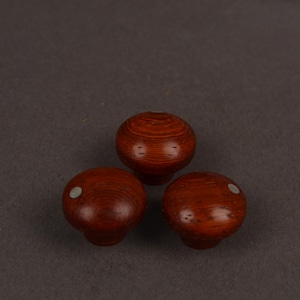 May include: Three round, reddish-brown wooden knobs with a smooth, polished finish. Each knob features a visible wood grain pattern and a small, circular, light-coloured inlay positioned off-centre on the top surface of two of the knobs.