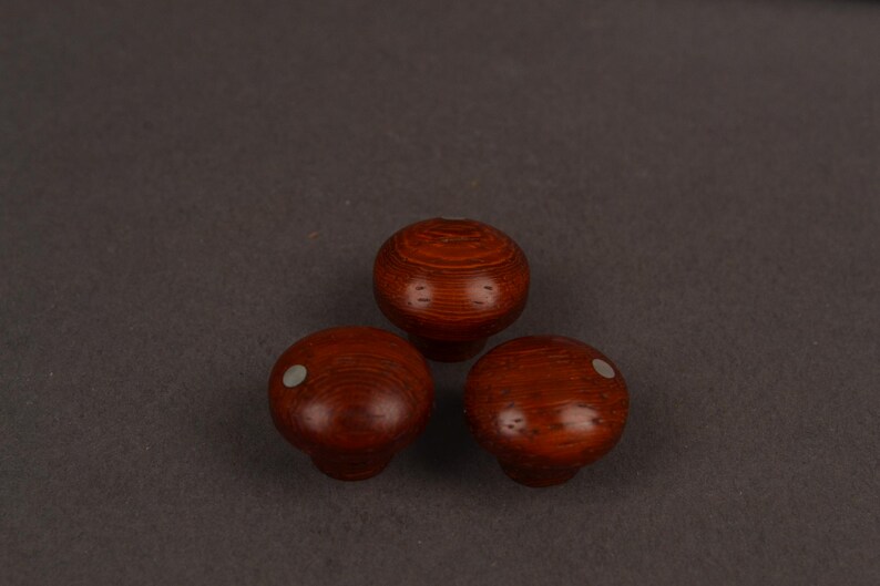 May include: Three round, reddish-brown wooden knobs with a smooth, polished finish. Each knob features a visible wood grain pattern and a small, circular, light-coloured inlay positioned off-centre on the top surface of two of the knobs.