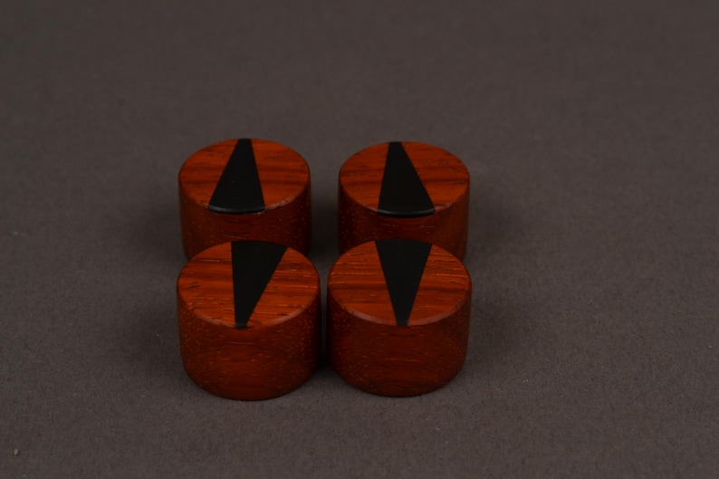 Peut inclure: Quatre objets cylindriques en bois de couleur brun rouge&acirc;tre. Chacun pr&eacute;sente une incrustation triangulaire noire sur la surface sup&eacute;rieure. Ils sont dispos&eacute;s en carr&eacute; sur un fond neutre.