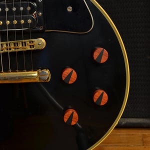 Peut inclure: Gros plan sur une guitare &eacute;lectrique noire avec des &eacute;l&eacute;ments dor&eacute;s et des boutons en bois. La guitare a une finition brillante et une bordure cr&egrave;me. Les boutons sont brun-rouge avec une incrustation noire.