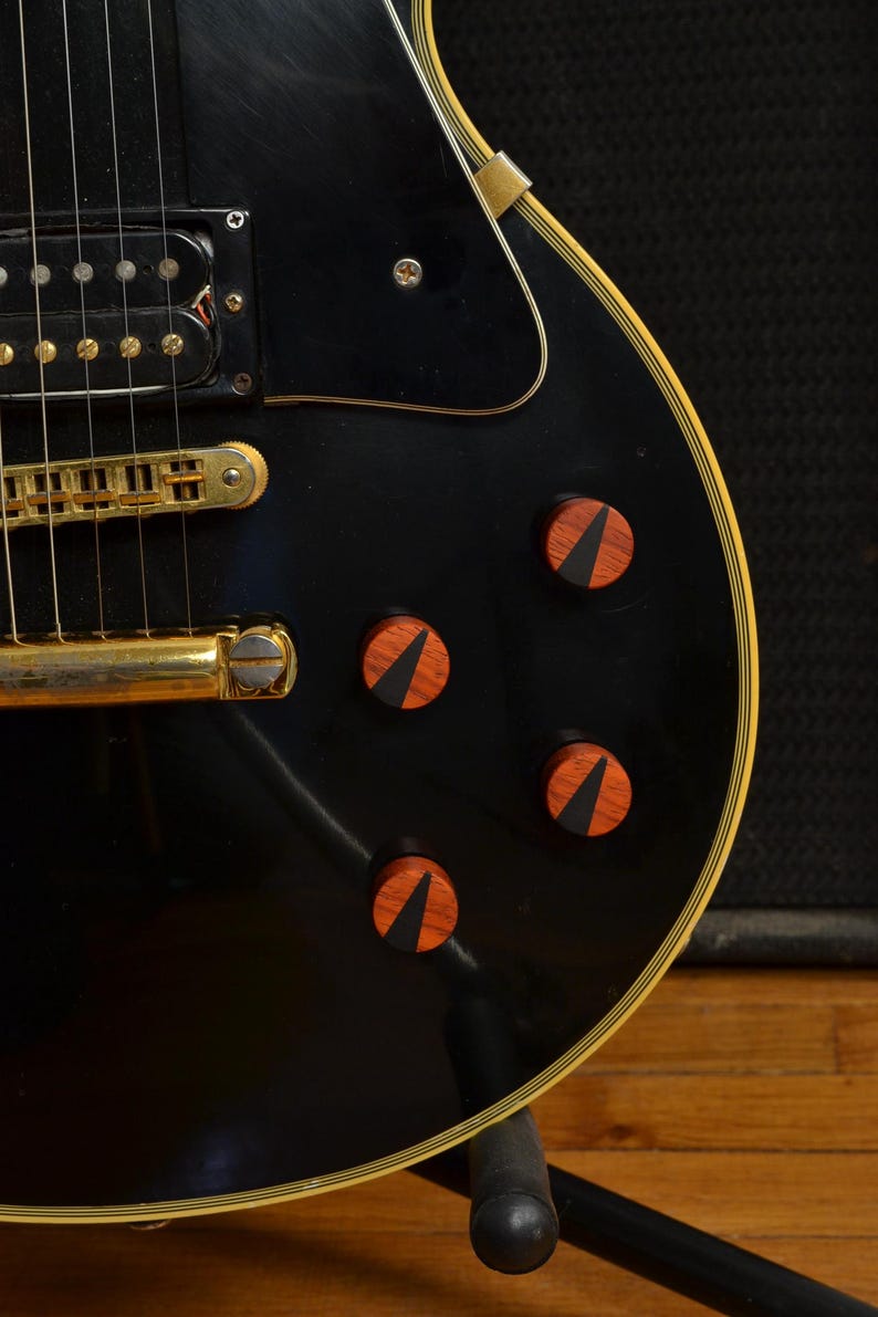 Peut inclure: Gros plan sur une guitare &eacute;lectrique noire avec des &eacute;l&eacute;ments dor&eacute;s et des boutons en bois. La guitare a une finition brillante et une bordure cr&egrave;me. Les boutons sont brun-rouge avec une incrustation noire.