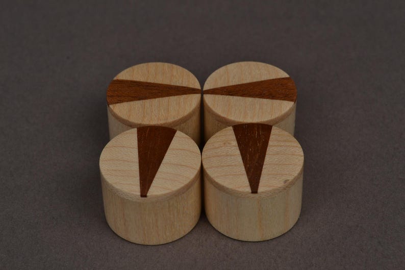 May include: Four small, cylindrical wooden boxes with light wood bodies and dark brown inlaid geometric designs on the lids. The boxes are arranged in a square formation against a neutral background.