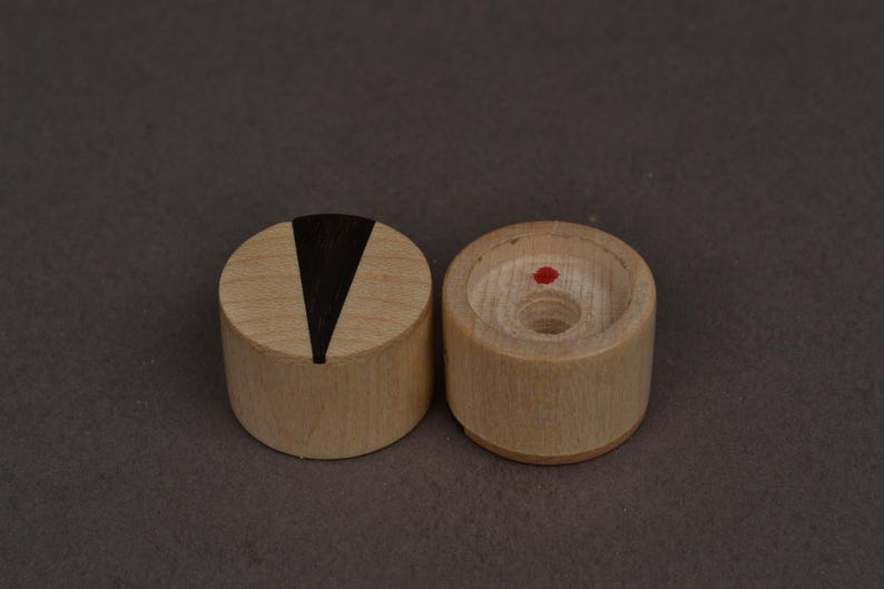 Op de afbeelding: Twee kleine, cilindervormige houten objecten. E&eacute;n heeft een zwarte V-vormige inleg op de bovenkant, de andere heeft een rode stip in het midden. Beide zijn gemaakt van lichtgekleurd hout en liggen op een donkergrijze ondergrond. Dit kunnen decoratieve items of onderdelen van een grotere set zijn.