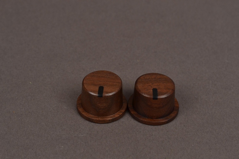May include: Two cylindrical wooden knobs with a dark brown finish and a visible wood grain pattern. Each knob features a flat top surface with a single vertical black indicator line running from the center toward the outer edge of the top.