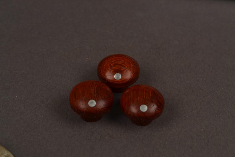 May include: Three round wooden knobs with a polished reddish-brown finish and visible wood grain patterns. Each knob features a small, circular, silver-coloured metal inlay centred on the top surface, providing a contrast to the dark wood.