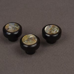 May include: Three black cabinet knobs with a round, iridescent inlay. The knobs have a dark, glossy finish and a classic, rounded shape. The inlay features a mix of colors, including gold, green, and white.