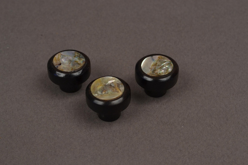 May include: Three black cabinet knobs with a round, iridescent inlay. The knobs have a dark, glossy finish and a classic, rounded shape. The inlay features a mix of colors, including gold, green, and white.
