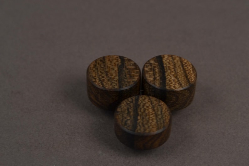Pode incluir: Tr&ecirc;s bot&otilde;es circulares de madeira com um padr&atilde;o de veios em castanho escuro e preto. Cada bot&atilde;o tem uma forma cil&iacute;ndrica lisa com uma superf&iacute;cie superior plana e uma textura natural vis&iacute;vel, exibindo os distintos veios escuros da madeira.