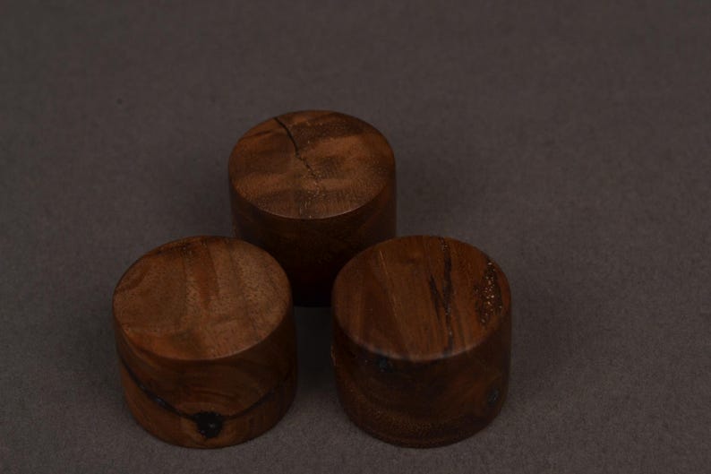 K&ouml;nnte beinhalten: Drei zylindrische Holzobjekte in einem satten Braunton mit sichtbarer Holzmaserung. Die Objekte sind in einem dreieckigen Muster vor einem neutralen Hintergrund angeordnet, wodurch ihre nat&uuml;rliche Textur und Form hervorgehoben werden.