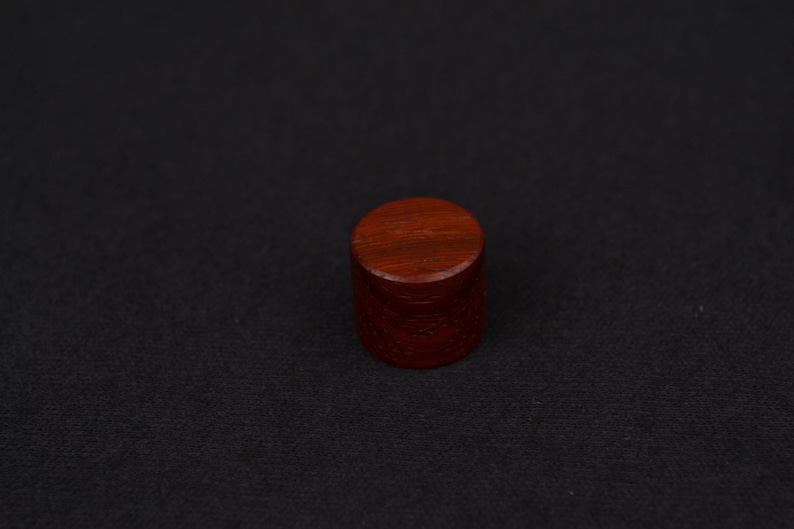 以下が含まれることがあります： 濃い赤褐色の無垢材で作られた円筒形の物体。上面と側面は滑らかで、暗い背景に配置されています。装飾要素またはより大きなアイテムのコンポーネントである可能性があります。