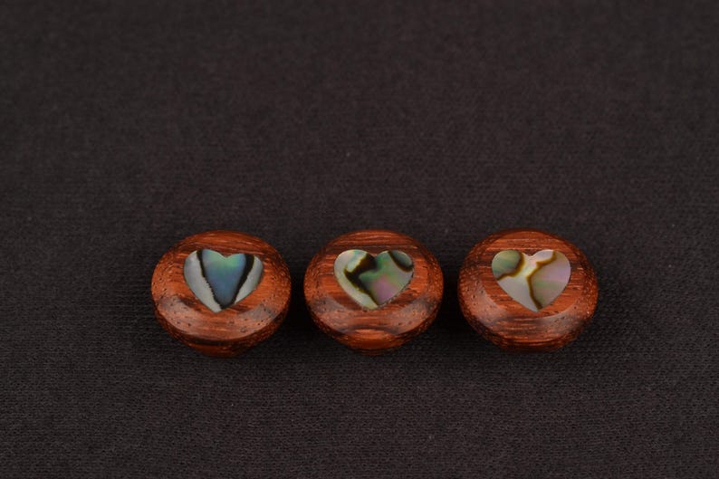 May include: Three round, wooden decorative buttons with a heart-shaped inlay of iridescent abalone shell. The buttons are a reddish-brown colour and are set against a dark background.