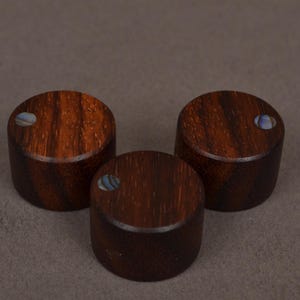 May include: Three cylindrical wooden knobs with a rich, dark brown grain pattern. Each knob features a small, iridescent inlay. The knobs are arranged in a triangular formation against a neutral background.