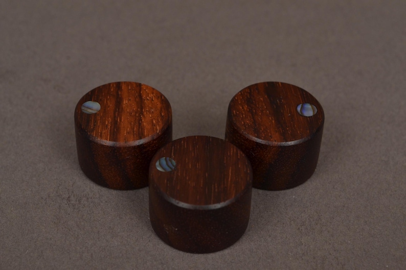 May include: Three cylindrical wooden knobs with a rich, dark brown grain pattern. Each knob features a small, iridescent inlay. The knobs are arranged in a triangular formation against a neutral background.