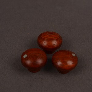 May include: Three round wooden knobs with a polished reddish-brown finish and visible wood grain. Two of the knobs feature a small, circular, light-coloured inlay on the top surface, while the third knob is plain with a smooth, rounded top.
