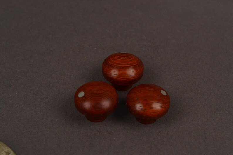May include: Three round wooden knobs with a polished reddish-brown finish and visible wood grain. Two of the knobs feature a small, circular, light-coloured inlay on the top surface, while the third knob is plain with a smooth, rounded top.