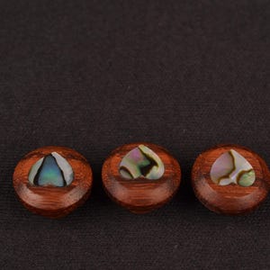 May include: Three round, wooden decorative buttons with a heart-shaped abalone inlay. The buttons are a rich brown colour, and the abalone has iridescent hues of blue, green, and pink. The buttons are arranged in a row against a dark background.