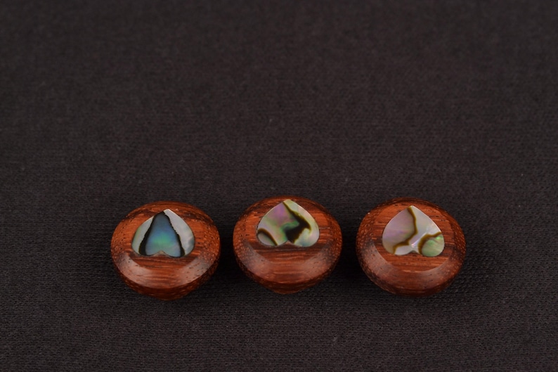 May include: Three round, wooden decorative buttons with a heart-shaped abalone inlay. The buttons are a rich brown colour, and the abalone has iridescent hues of blue, green, and pink. The buttons are arranged in a row against a dark background.