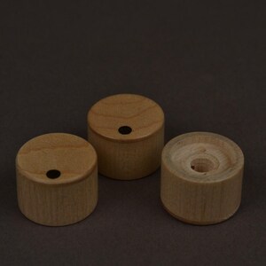 May include: Three small, cylindrical wooden objects. Two have a small, dark hole in the center of the top surface. The third has a larger, central hole. All are light brown and sit on a dark brown background.