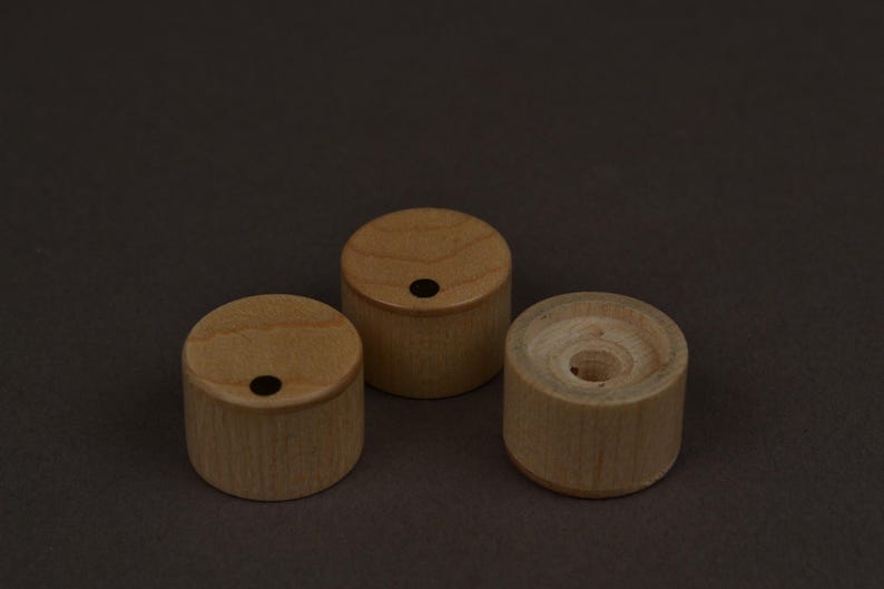 May include: Three small, cylindrical wooden objects. Two have a small, dark hole in the center of the top surface. The third has a larger, central hole. All are light brown and sit on a dark brown background.