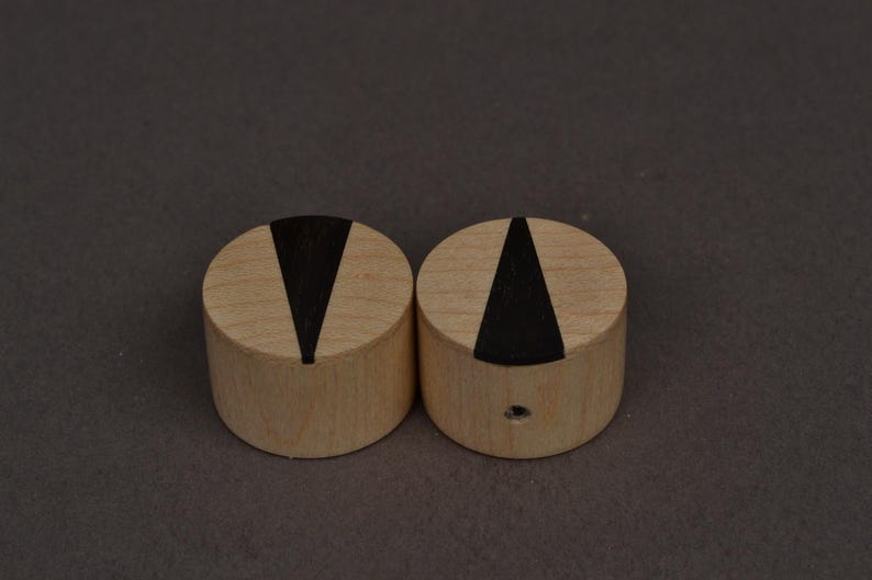 Op de afbeelding: Twee cilindrische houten objecten met een lichtbruine afwerking. Elk heeft een zwarte, driehoekige inleg op het bovenoppervlak. De objecten staan naast elkaar tegen een donkergrijze achtergrond. E&eacute;n object heeft een klein gaatje aan de zijkant.