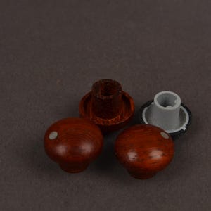 May include: Two round knobs with a reddish-brown wood grain finish and a small white dot on the top surface. Each knob features a circular base with a central mounting hole, one showing a dark brown interior and the other a light grey plastic insert.