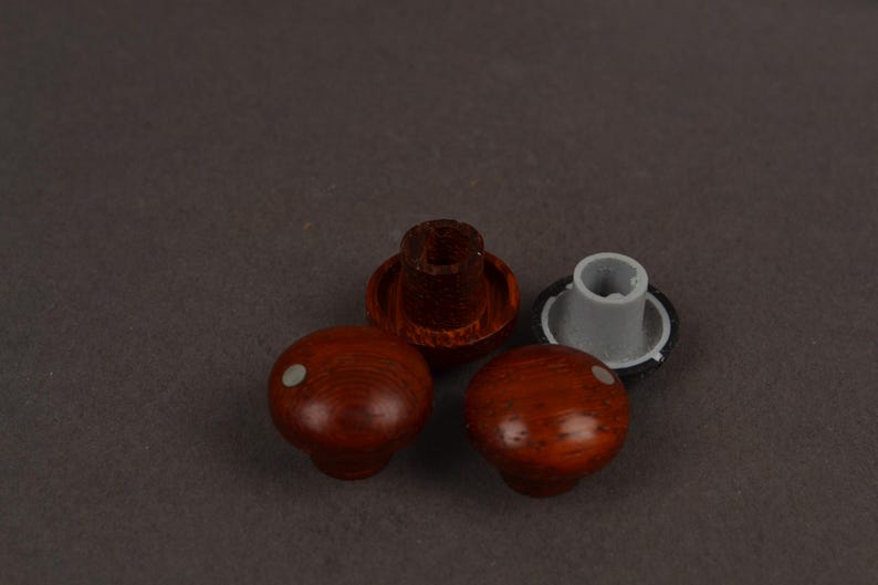 May include: Two round knobs with a reddish-brown wood grain finish and a small white dot on the top surface. Each knob features a circular base with a central mounting hole, one showing a dark brown interior and the other a light grey plastic insert.