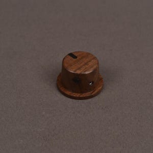 May include: A cylindrical dark brown wooden control knob featuring a visible wood grain pattern, a flat top surface with a single thin black indicator line, and a small metallic set screw hole located on the side of the lower base section.
