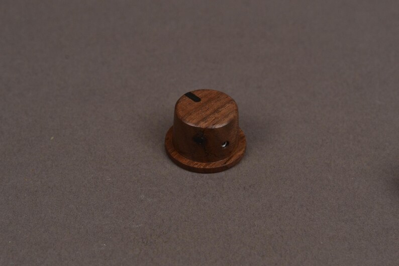 May include: A cylindrical dark brown wooden control knob featuring a visible wood grain pattern, a flat top surface with a single thin black indicator line, and a small metallic set screw hole located on the side of the lower base section.