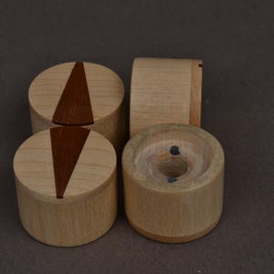 May include: Four wooden objects. Two are cylindrical with a dark brown triangular inlay on top. One is a cylinder with a curved side. The last is a cylinder with a hole in the center. The objects are made of light-colored wood.