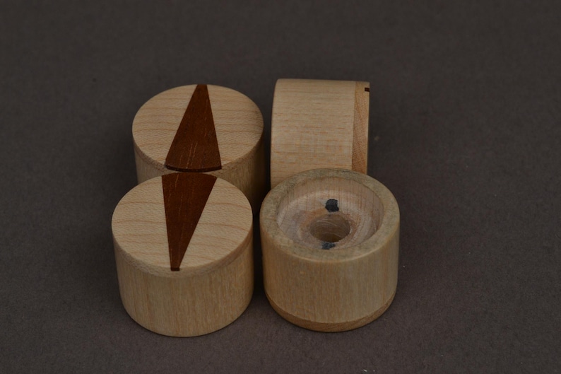 May include: Four wooden objects. Two are cylindrical with a dark brown triangular inlay on top. One is a cylinder with a curved side. The last is a cylinder with a hole in the center. The objects are made of light-colored wood.
