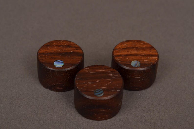 May include: Three wooden knobs with a rich, dark brown grain. Each knob has a small, round inlay of iridescent blue and white material. The knobs are cylindrical and appear to be handmade.