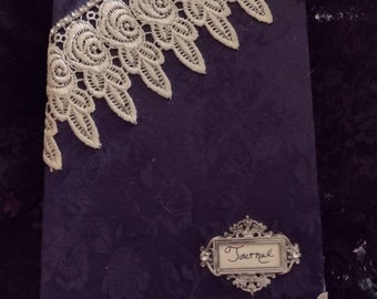 May include: A dark blue journal with a floral pattern, adorned with white lace trim and silver corner protectors. The journal features a decorative silver label that reads "Journal".