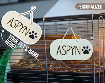 May include: A personalized wooden sign with the name "Aspyn" and a paw print, hanging on a cage. The sign is white with black lettering and a black paw print. The sign is for a pet and can be customized with any name.
