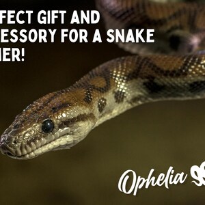 May include: A close-up of a brown and black snake with a white eye. The snake is curled up and has a textured skin. The text "PERFECT GIFT AND ACCESSORY FOR A SNAKE OWNER!" is written above the snake. The text "Ophelia SS" is written below the snake.