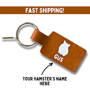 May include: A brown leather keychain with a white hamster silhouette and the name "Gus" printed on it. The keychain has a silver key ring.
