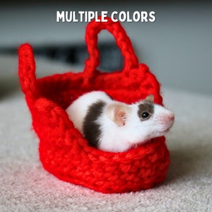 May include: A small white and brown mouse sitting in a red crocheted basket. The basket has a handle and is made of yarn.