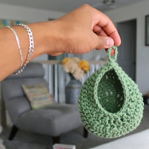 May include: A green crocheted hanging basket with a loop at the top. The basket is made with a textured stitch and has a rounded shape.