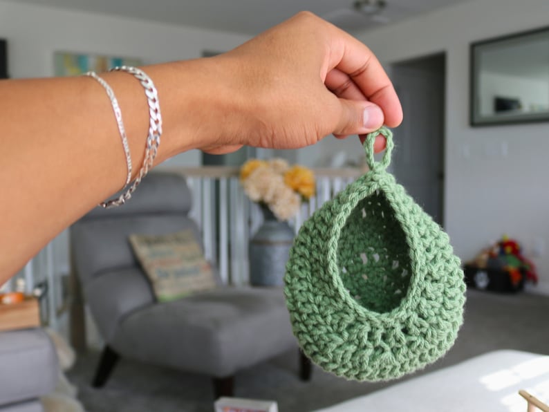 May include: A green crocheted hanging basket with a loop at the top. The basket is made with a textured stitch and has a rounded shape.