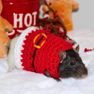 May include: A black rat wearing a red and white crocheted Santa Claus costume. The rat is lying on a white fluffy surface.