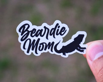Puede incluir: Una pegatina blanca con las palabras "Beardie Mom" en fuente negra, acompañada de una silueta negra de un dragón barbudo. La pegatina tiene un borde festoneado.