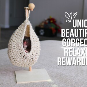 May include: A small hamster resting in a beige crocheted hanging hammock. The hammock is suspended from a wooden stand with a small wooden ball on top.