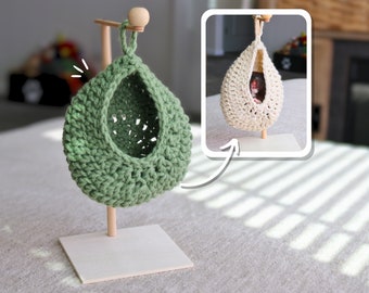 May include: A green crocheted hanging hammock for small pets, like hamsters or gerbils, is shown on a wooden stand. The hammock is suspended from the stand by a wooden dowel. The hammock is made of a soft, crocheted material and has a round shape. The stand is made of wood and has a square base.