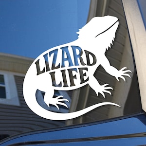 May include: White vinyl decal of a lizard with the words "Lizard Life" in the center. The decal is applied to a car window, with a house and blue sky visible in the background. The lizard is a bearded dragon.