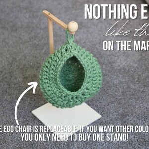 May include: A green crocheted egg chair hanging from a wooden stand. The stand is white and has a round wooden knob at the top. The text "NOTHING ELSE like this! ON THE MARKET!" is written in black on a white background. The text "THE EGG CHAIR IS REPLACEABLE IF YOU WANT OTHER COLORS! YOU ONLY NEED TO BUY ONE STAND!" is written in black on a white background.