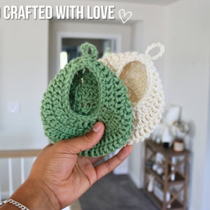 May include: Two crocheted pot holders, one green and one white, hanging from a loop. The green pot holder is being held in a hand.