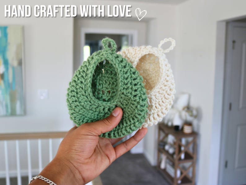 May include: Two crocheted pot holders, one green and one white, hanging from a loop. The green pot holder is being held in a hand.