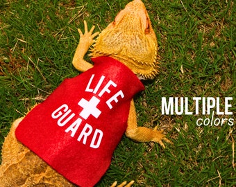 May include: A bearded dragon wearing a red lifeguard vest with white text that says "LIFE GUARD" and a white cross. The text "MULTIPLE colors" is written in white on a black background below the dragon.